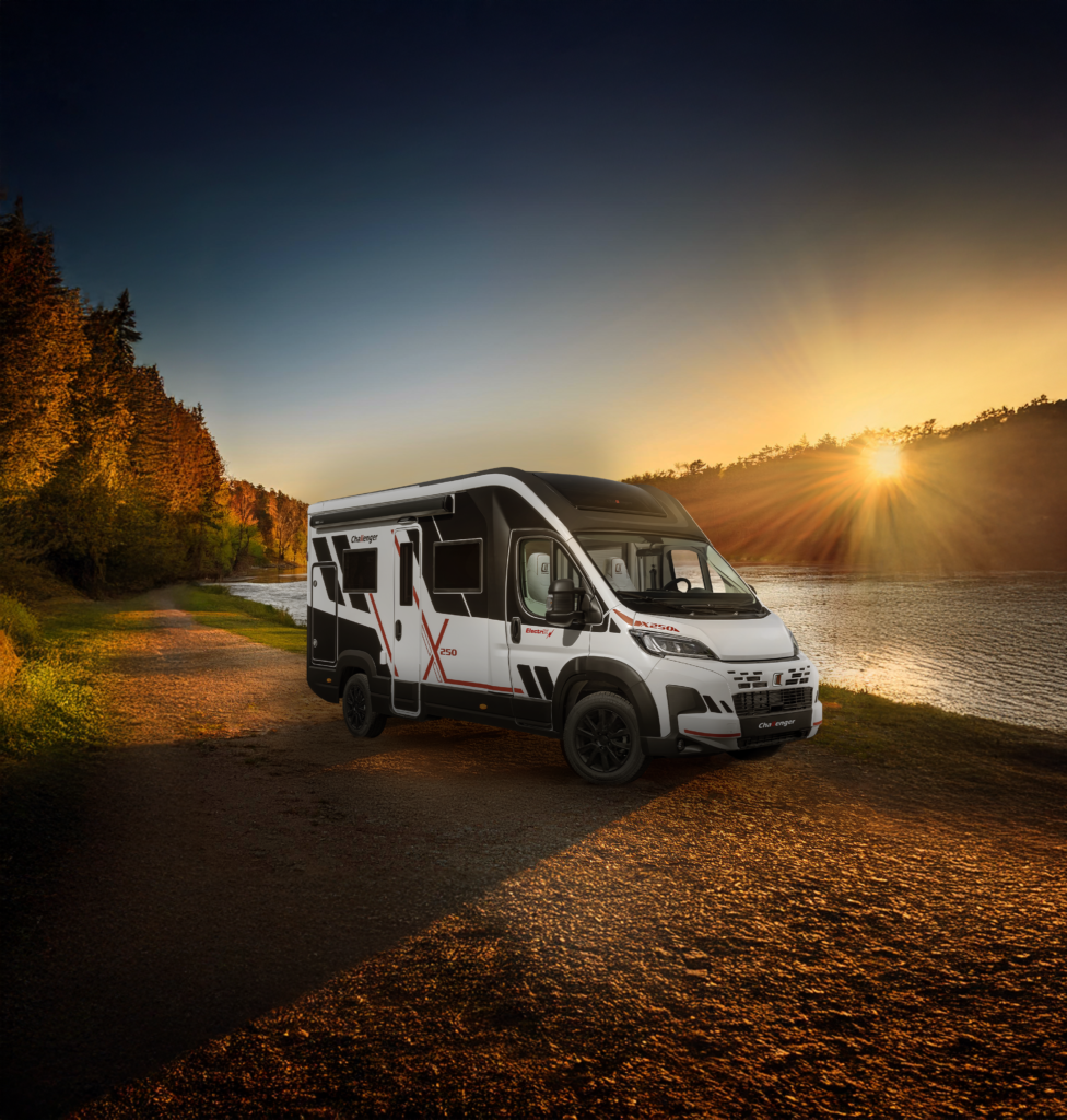 A white motorhome with black accents parked up on the waterside by a river. The surrounding backdrop is a sunset peaking through a hillside of trees on the opposite side of the river.