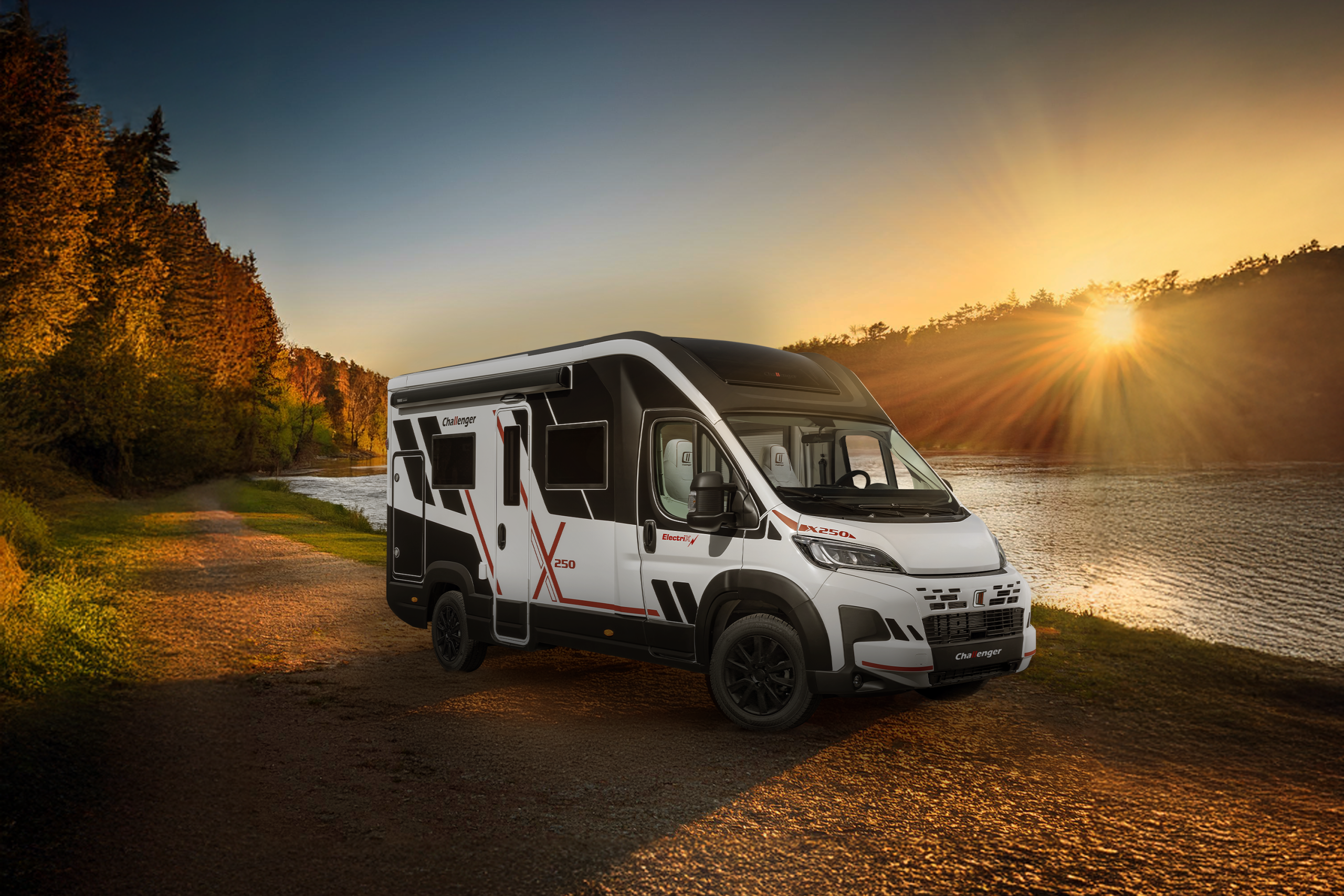 A white motorhome with black accents parked up on the waterside by a river. The surrounding backdrop is a sunset peaking through a hillside of trees on the opposite side of the river.